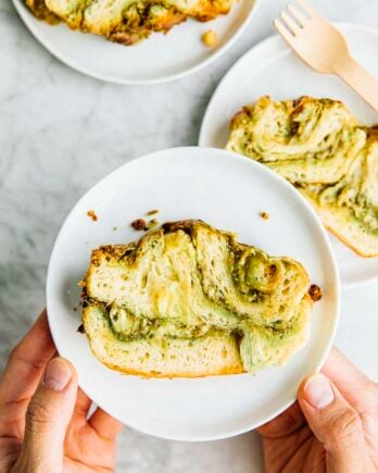 A photo of hands holding a slice of white chocolate pistachio babka on a white plate.