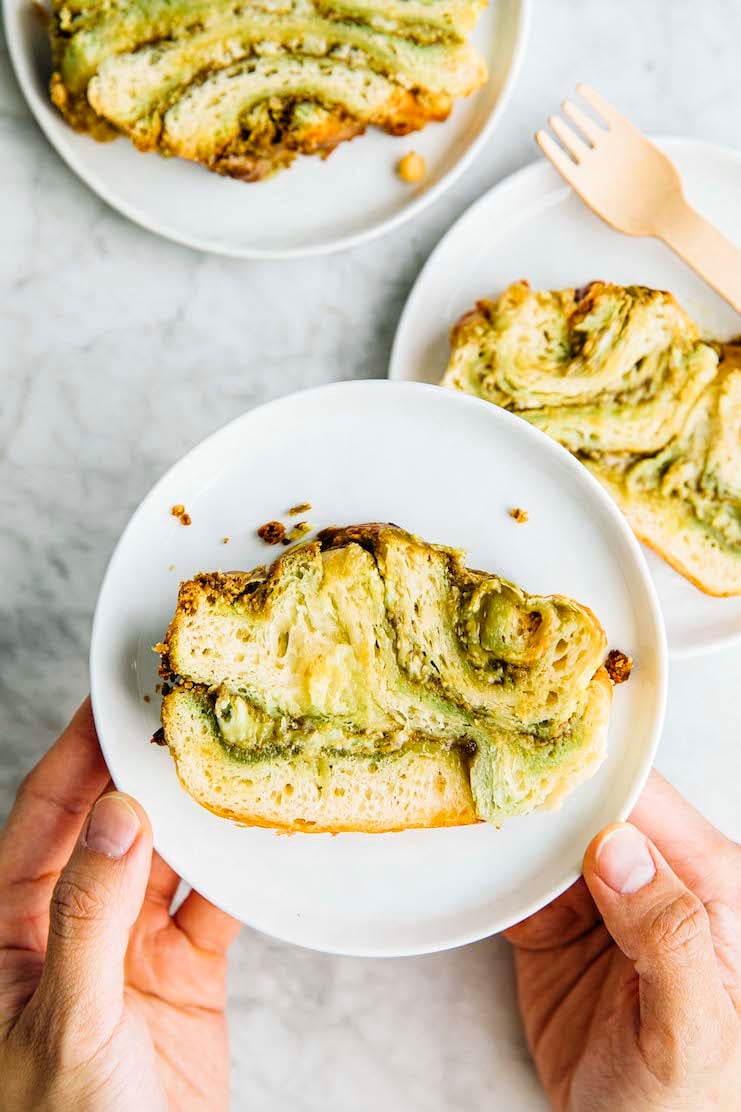 A photo of hands holding a slice of white chocolate pistachio babka on a white plate.