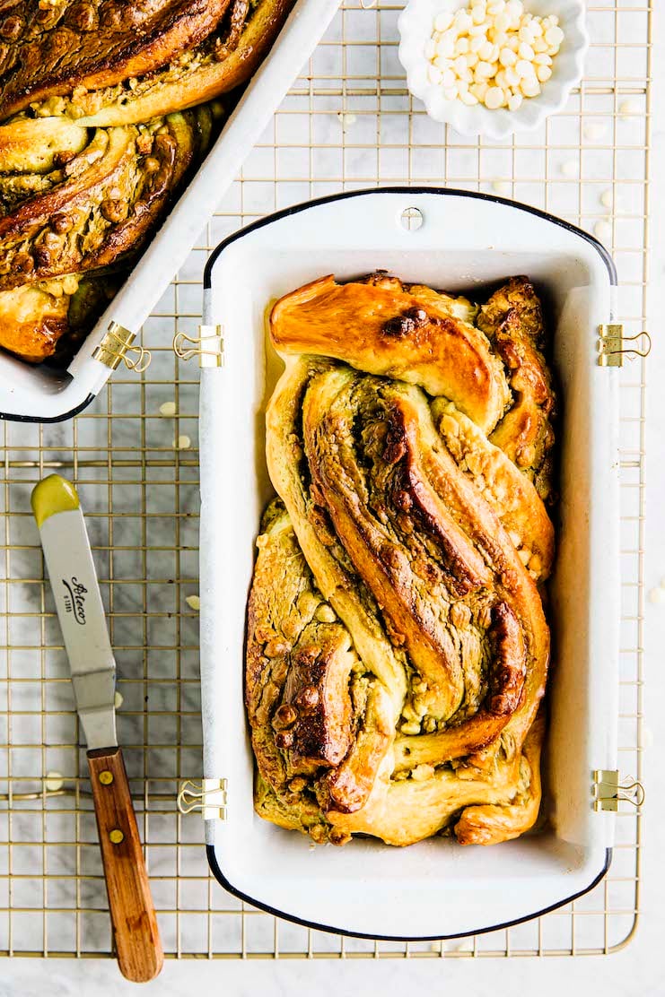 A photo of white chocolate pistachio babka in a pan on a wire rack next to an offset spatula. 