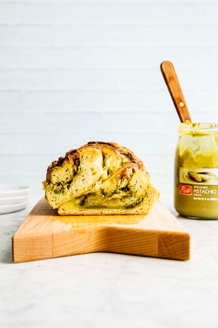 A photo of a white chocolate pistachio babka sliced showing off its swirly crumb next to a jar of pistachio cream. 