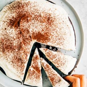 A photo of a chocolate cake with white frosting on a white plate with a hand reaching for a slice.