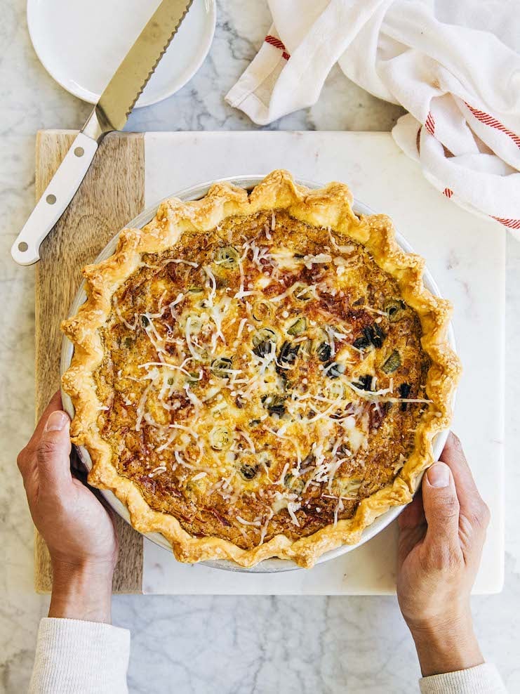 A picture of hands holding a quiche on a square marble platter.