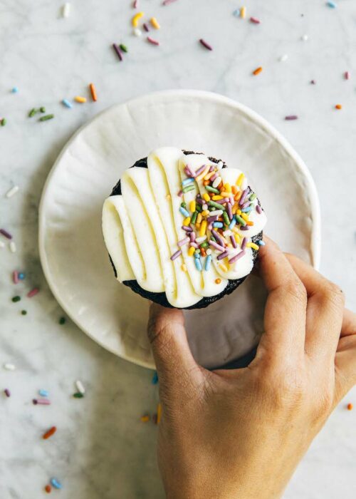 photo of hand reaching for chocolate cupcake on white plate with sprinkles