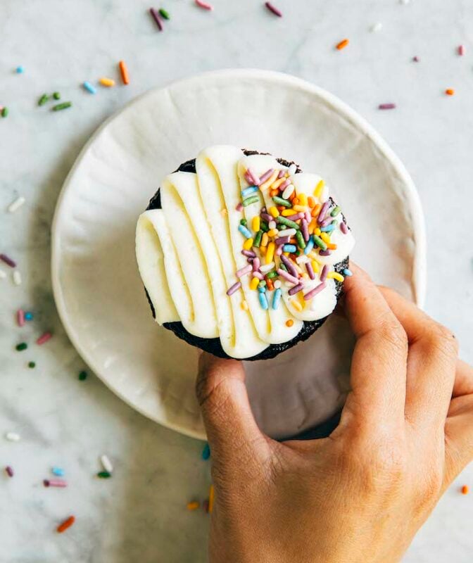 photo of hand reaching for chocolate cupcake on white plate with sprinkles