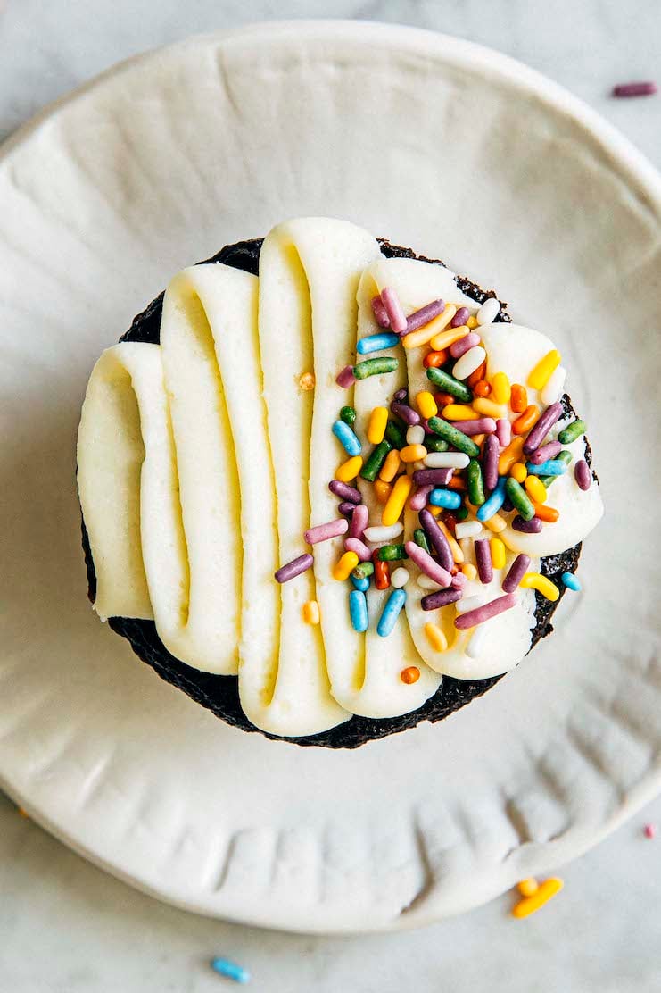 close up photo of chocolate cupcake with sprinkles and squiggle frosting on white plate