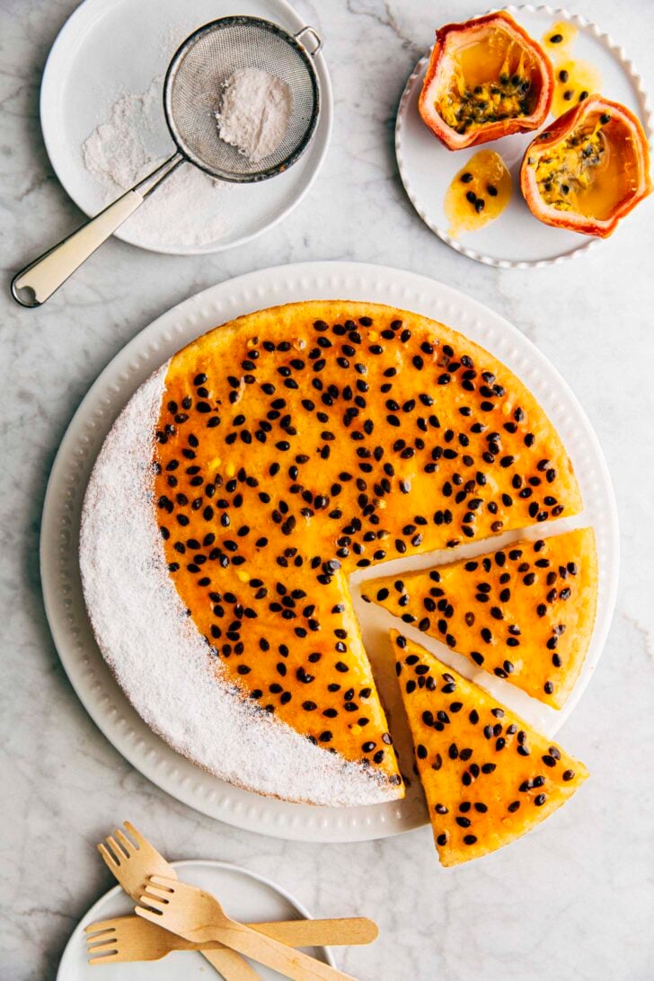 photo of passion fruit butter mochi cake sliced on a white plate surrounded by passion fruit and powdered sugar